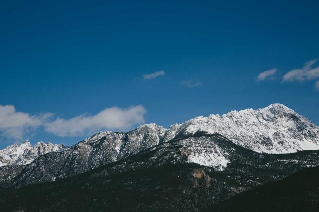 Schneebedeckte Berge unter einem klaren, blauen Himmel mit ein paar Wolken und baumbedeckten Hängen im Vordergrund.