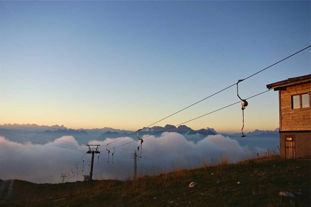 Ein Skilift auf einer grasbewachsenen Anhöhe bei Sonnenuntergang, mit Wolken darunter und Berggipfeln in der Ferne; rechts ist ein Holzgebäude zu sehen.