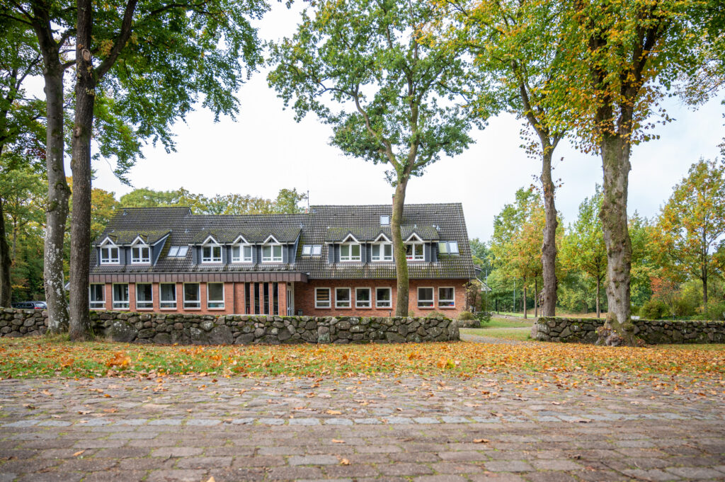 Ein großes Backsteinhaus mit vielen Fenstern ist von Bäumen und Steinmauern umgeben, auf dem Boden liegen an einem bewölkten Tag verstreute Blätter.
