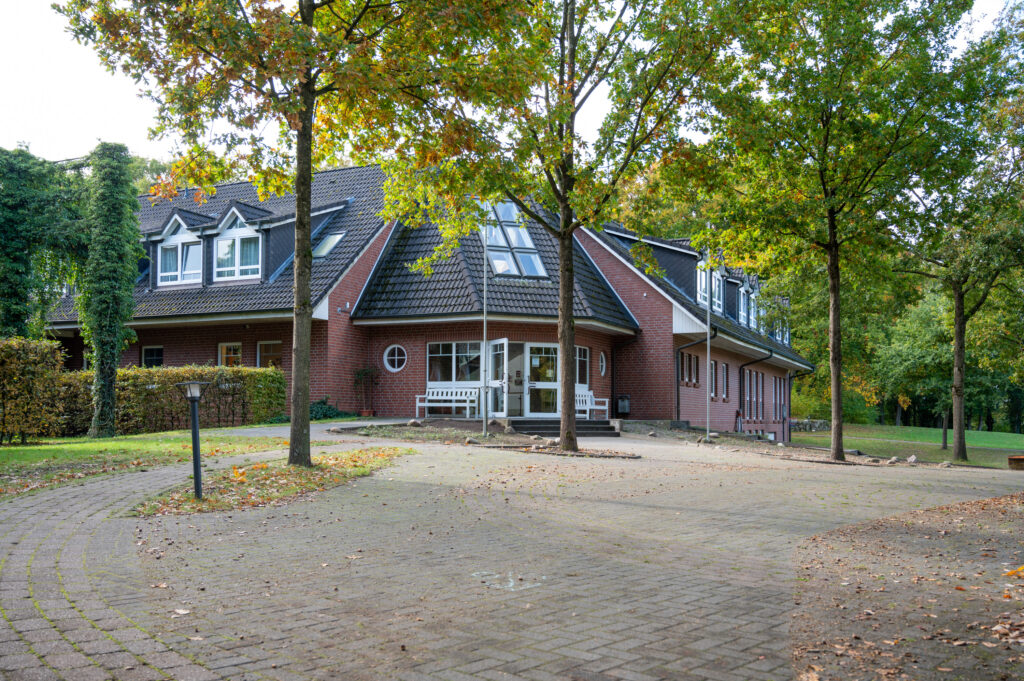 Ein großes Backsteinhaus mit einem dunklen Dach und vielen Fenstern ist an einem klaren Tag von Bäumen und einer gepflasterten Auffahrt umgeben.