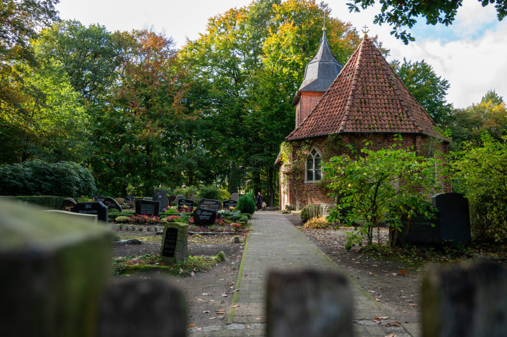 Eine kleine Backsteinkapelle mit einem steilen, roten Ziegeldach steht inmitten eines Friedhofs, umgeben von Grabsteinen, Bäumen und üppigem Grün unter einem teilweise bewölkten Himmel.